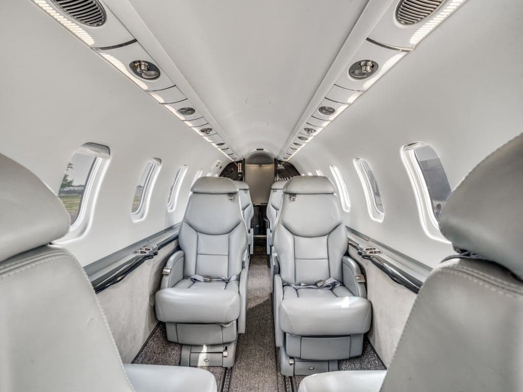 The interior cabin of a private jet, showing two rows of grey leather seats facing each other.