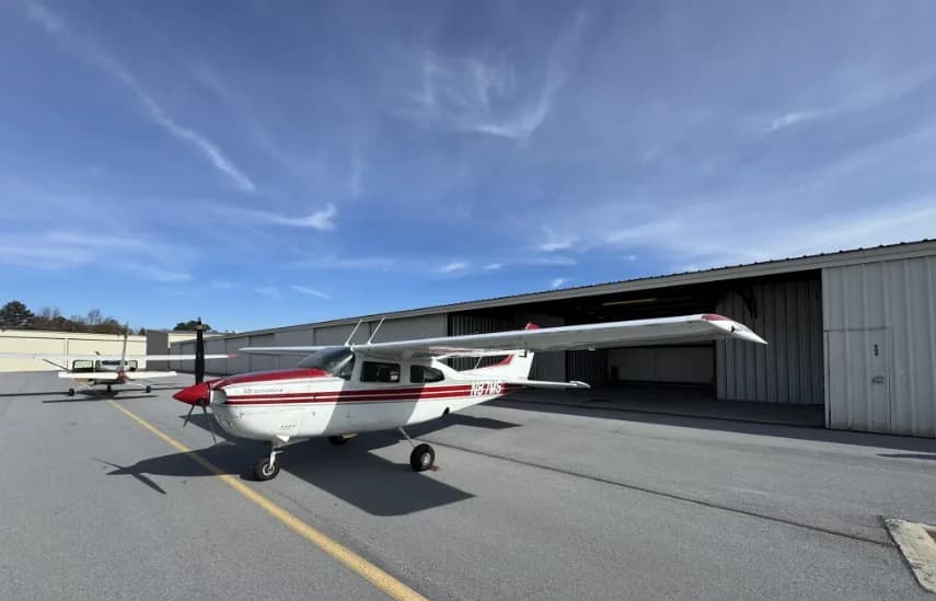 A white and red Cessna 182 Skylane (N87MS) parked on an airport tarmac next to hangars under a blue sky.