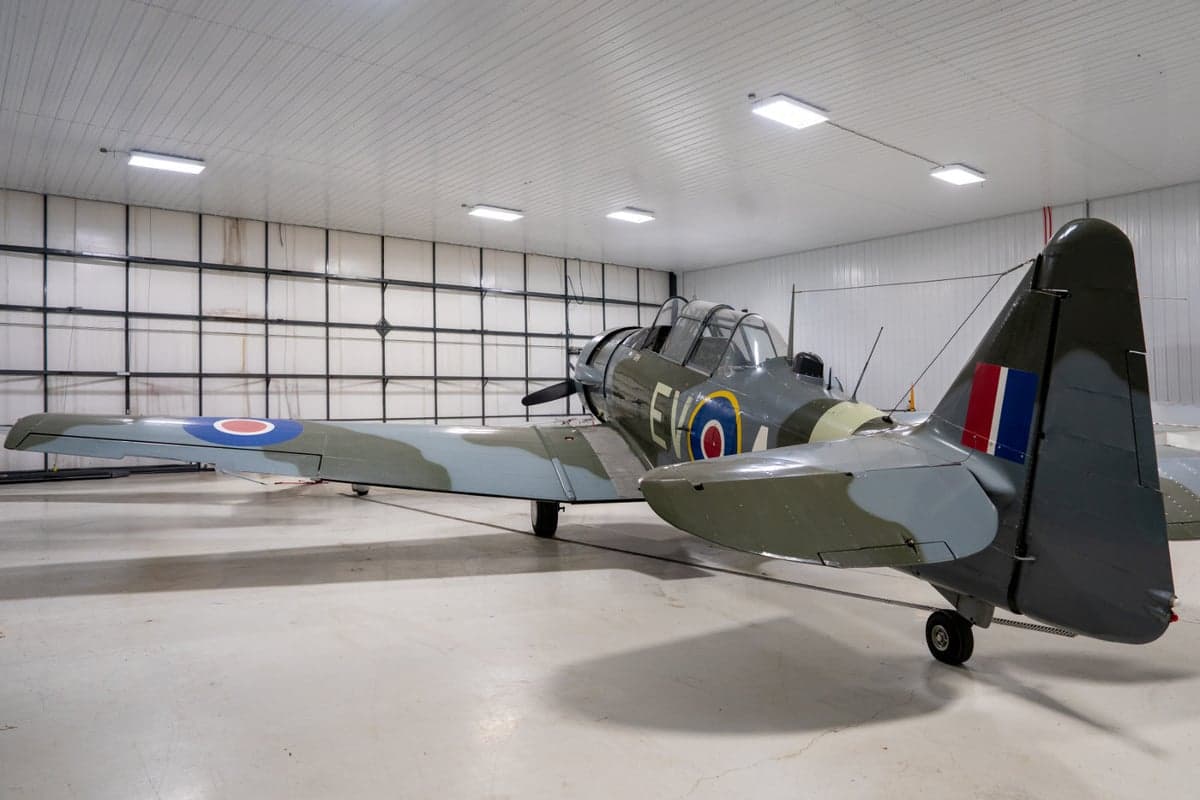 A three-quarter rear view of a vintage military trainer aircraft with a camouflage paint scheme, parked in a white hangar.