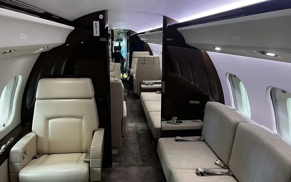 The interior of a private jet cabin showing cream-colored seats and a sofa, with dark wood paneling.