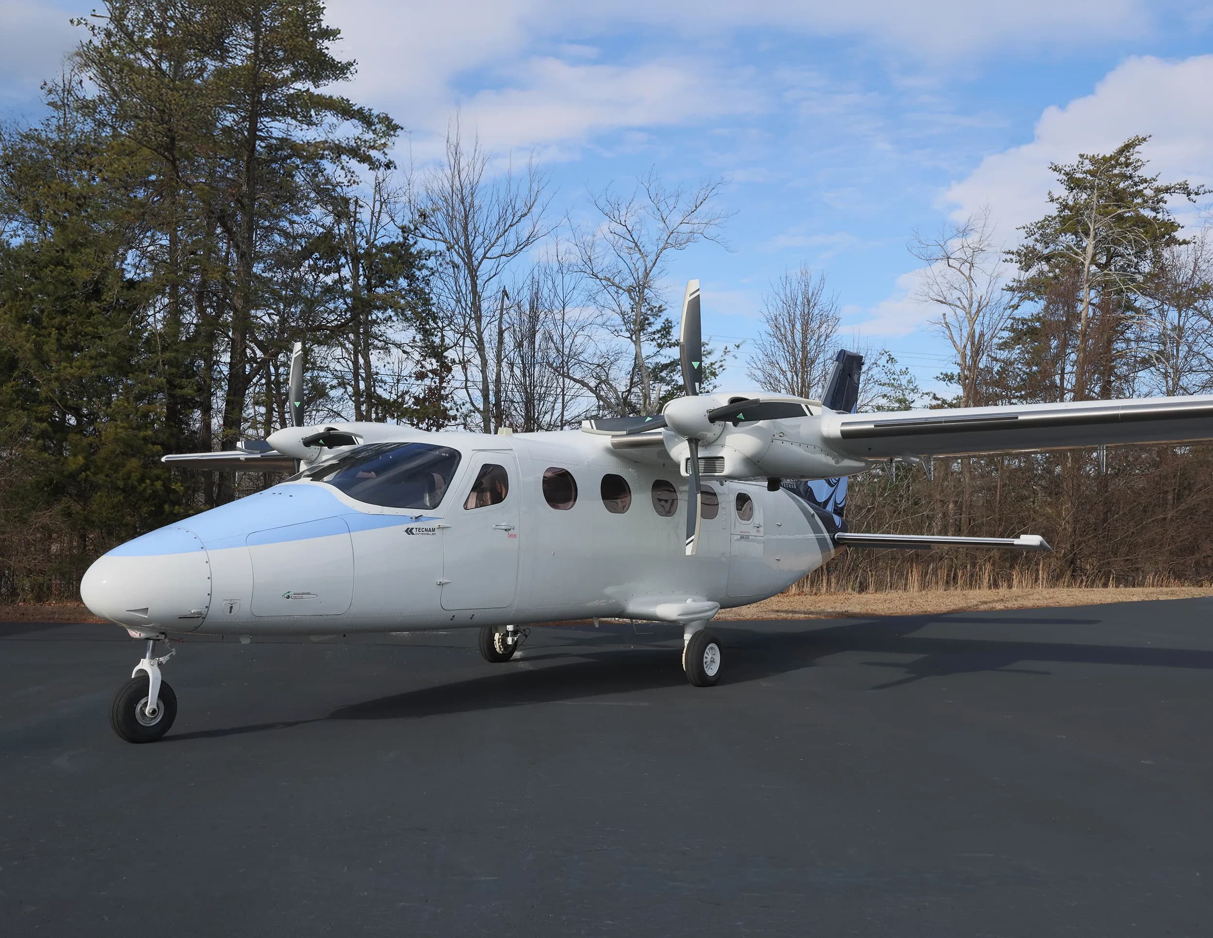 A white and blue twin-engine aircraft with a floral tail logo parked on an asphalt ramp, viewed from the front-right.
