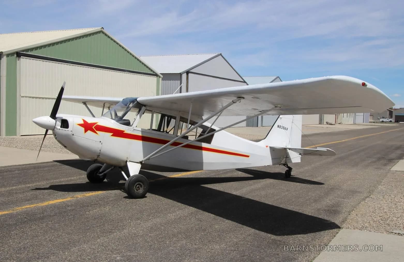 A white single-engine high-wing aircraft with red stripes and a red star on the cowl, parked on an asphalt taxiway with hangars in the background.