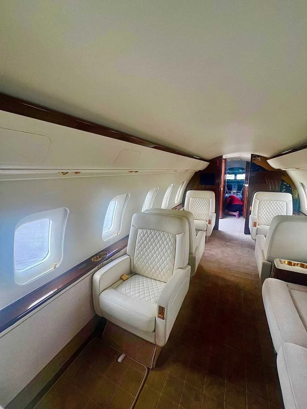 A view down the private jet cabin, showing quilted white leather seats, large windows, and dark wood accents.