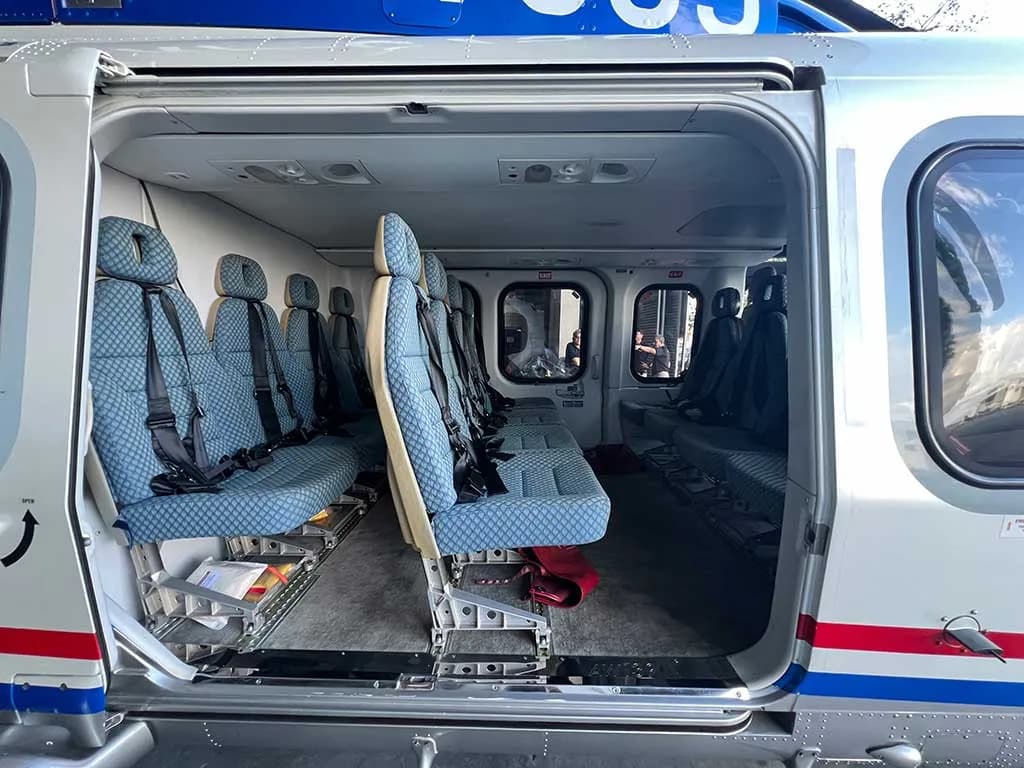 The open side door of a blue and white helicopter revealing rows of blue passenger seats inside a hangar.