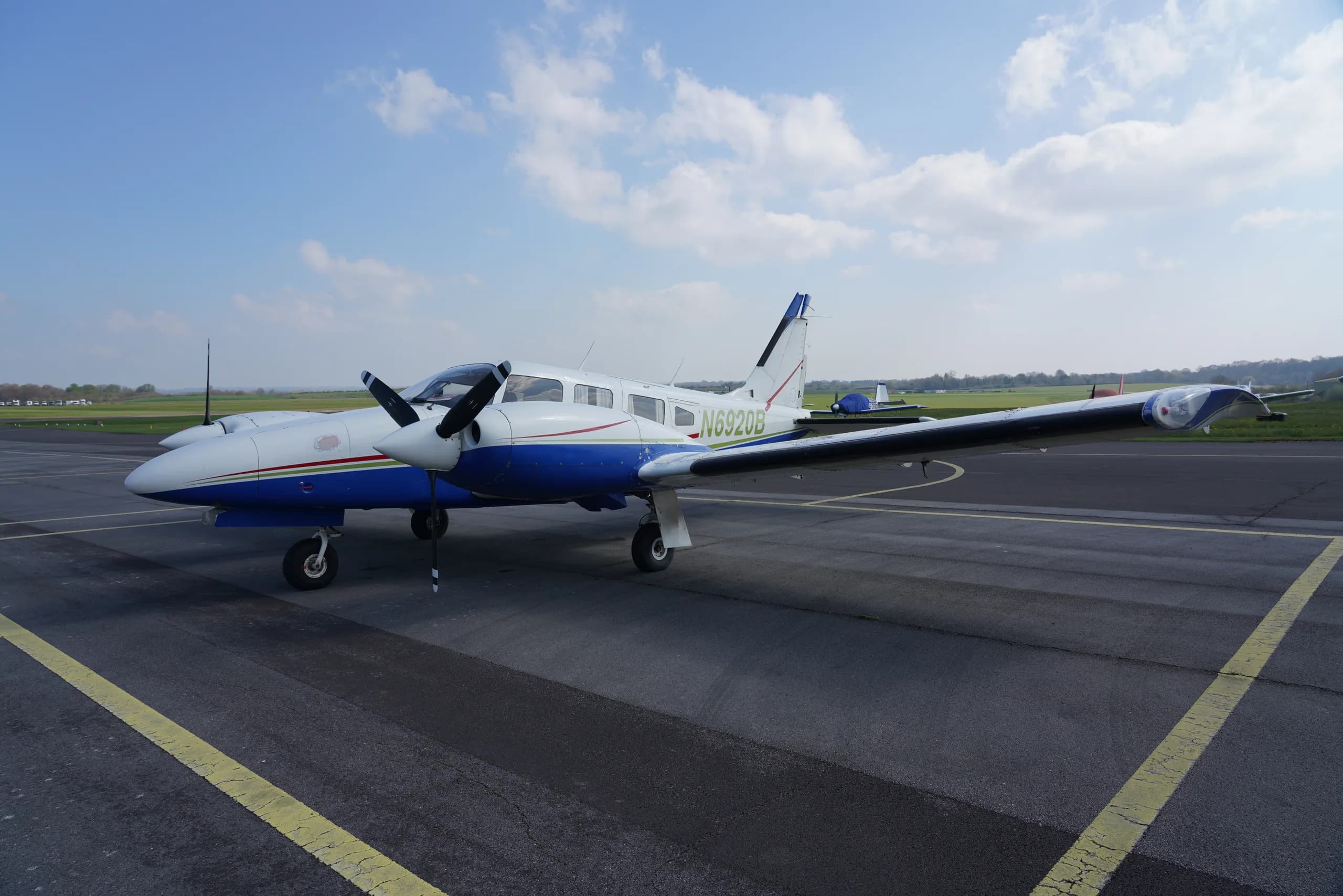 A white and blue twin-engine propeller aircraft with registration N6920B parked on an airport tarmac under a partly cloudy sky.