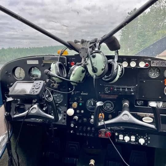 The cockpit of a floatplane shows the instrument panel, controls, and two headsets hanging.