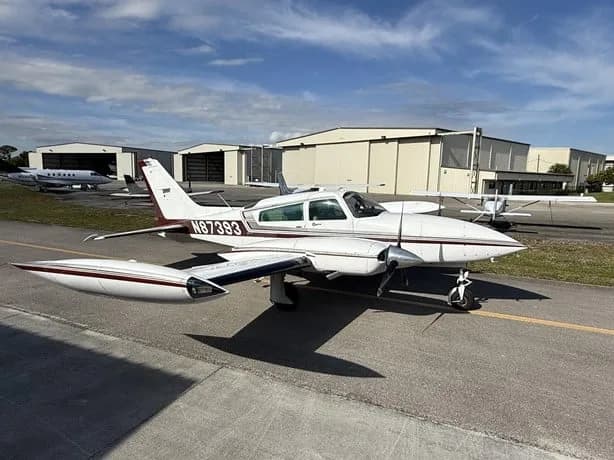 A white twin-engine aircraft with N87393 on the tail, parked on an airfield with hangars.