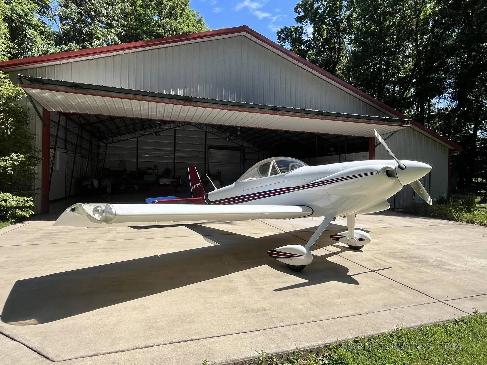 A white single-engine aircraft with red and blue stripes, parked on concrete in front of an open hangar, viewed from the front-right.