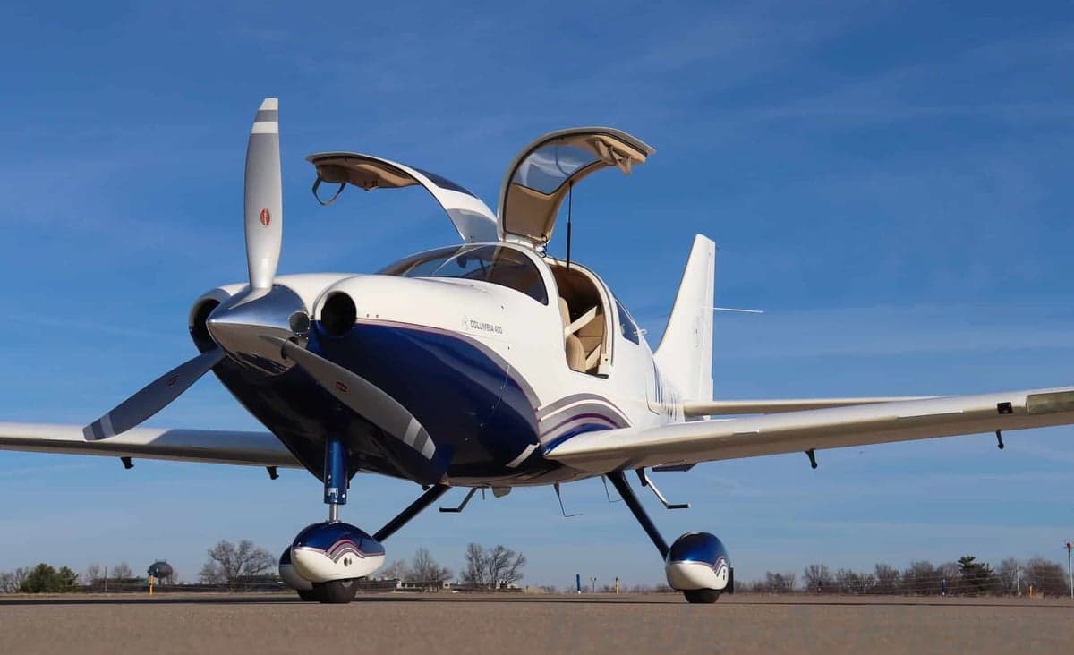 Low-angle front view of a white and blue Columbia 400 aircraft with its gull-wing doors open on an airfield.