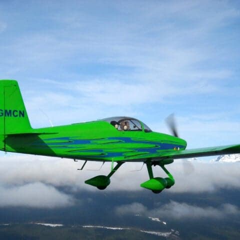 A green aircraft with blue accents in flight against a cloudy sky.