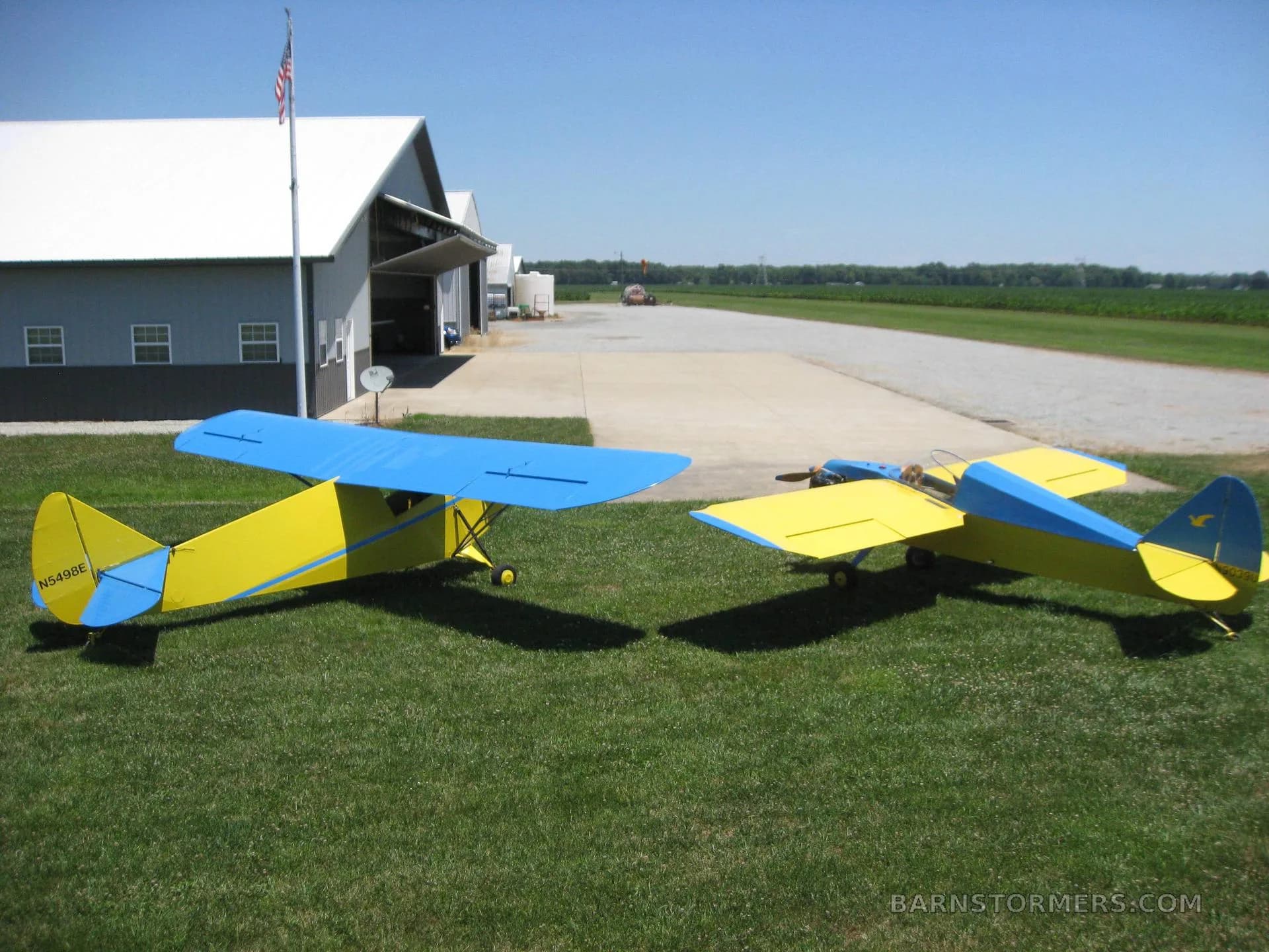 Two yellow and blue single-engine aircraft parked on grass near a hangar and runway.