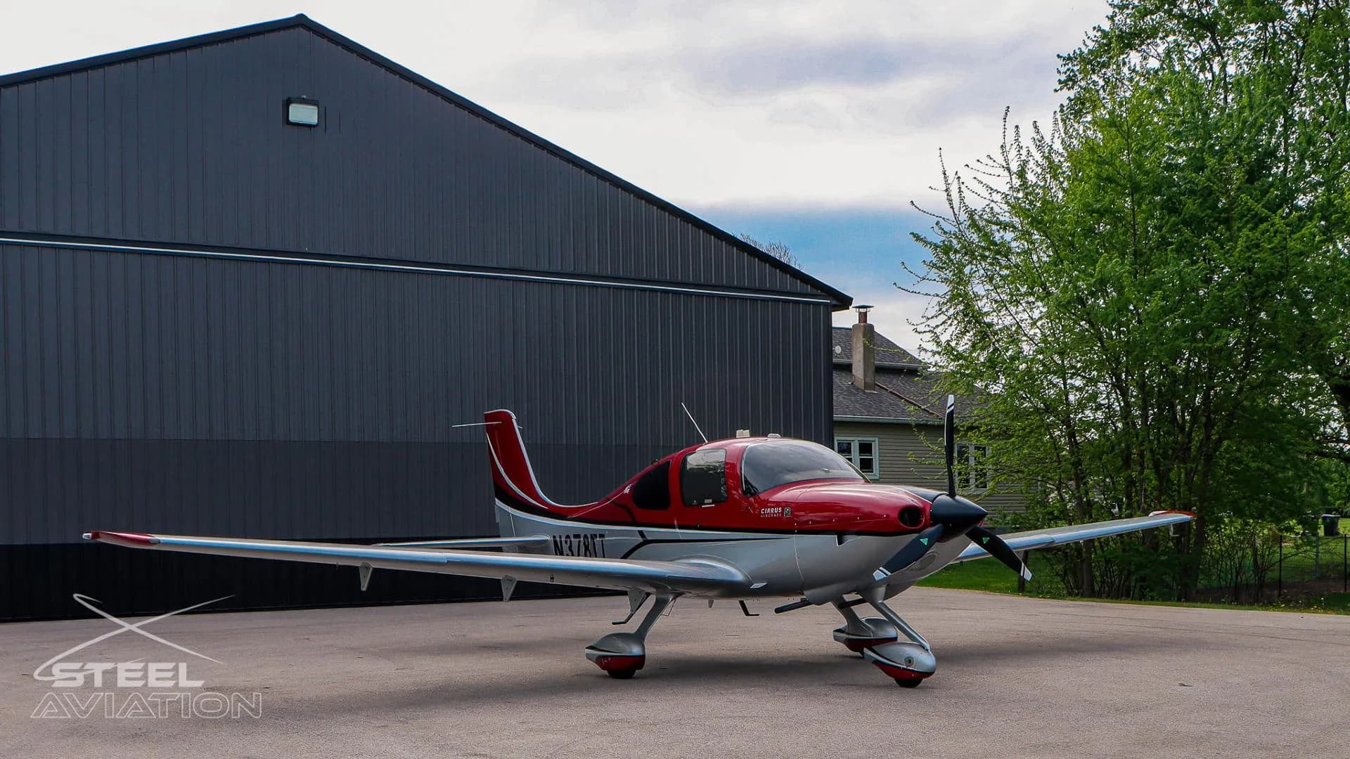 A red and white Cirrus SR22 aircraft with tail number N378TT is parked on an apron next to a dark gray hangar and green trees.