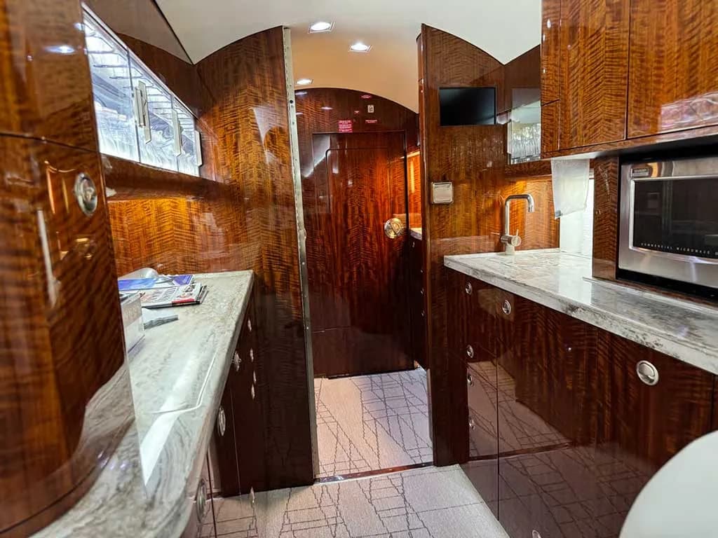 The aircraft galley features glossy dark wood cabinetry, granite countertops, a sink, and a microwave.