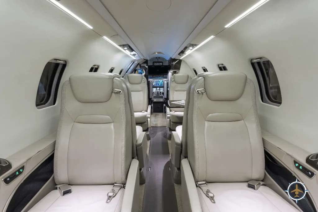 A view down the aisle of a private jet cabin with light-colored leather seats and overhead lighting.
