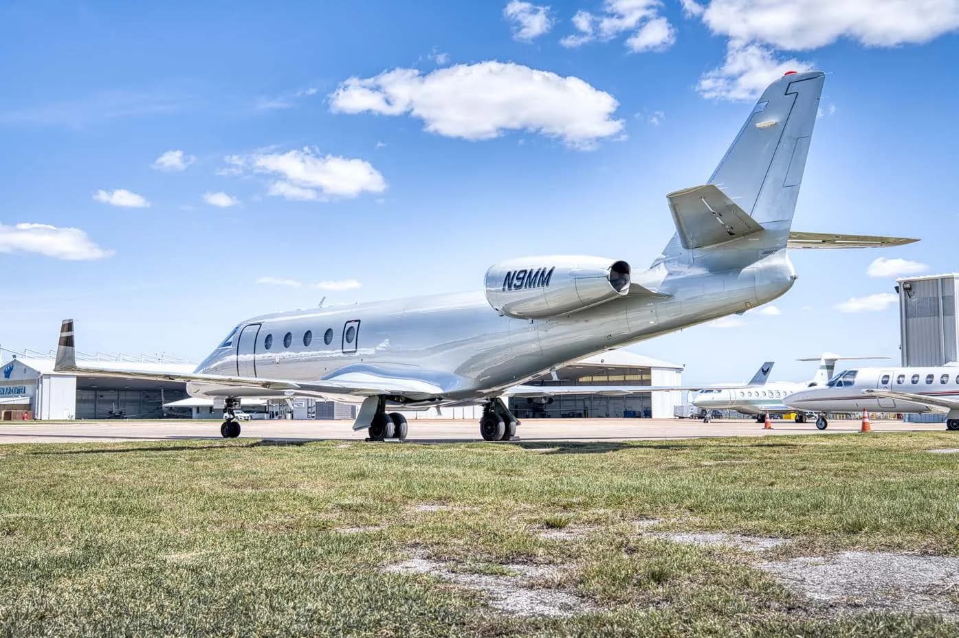 A silver business jet with registration N9MM parked on an airfield with other aircraft in the background.