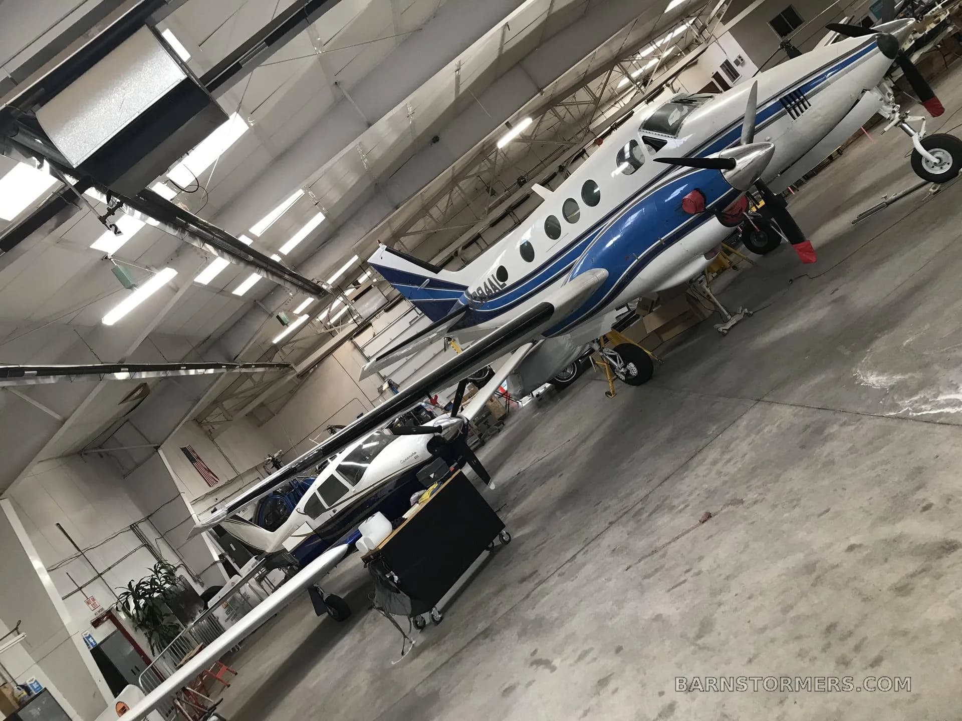 A white and blue twin-engine turboprop aircraft is parked in a hangar, with another aircraft partially visible behind it.