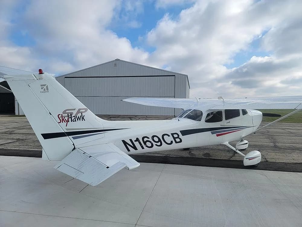 A rear-side view of the white Cessna Skyhawk N169CB with black and red stripes, parked on an airport ramp with a white hangar in the background.