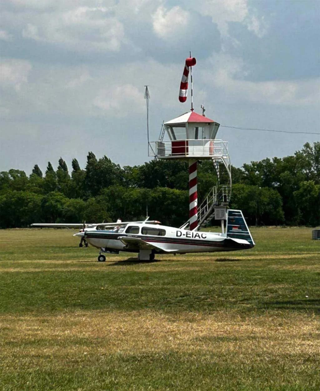 A Mooney M20J aircraft with a silver, blue, and red paint scheme parked on a grassy field with a small red and white striped control tower in the background.