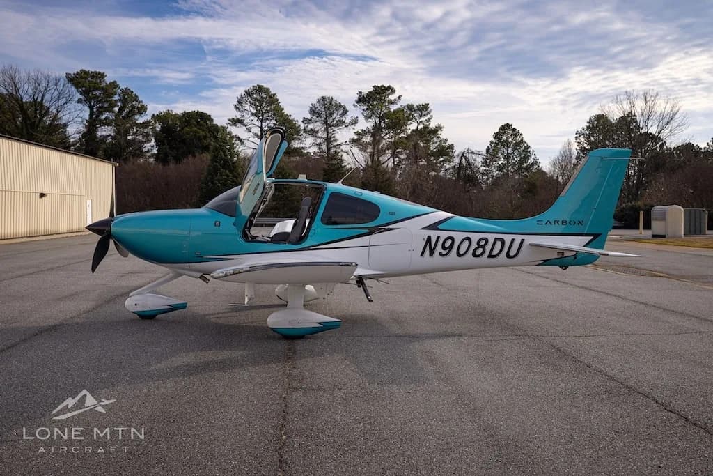 A side view of the teal and white Cirrus SR22 aircraft, N908DU, with its doors open, on an airfield.