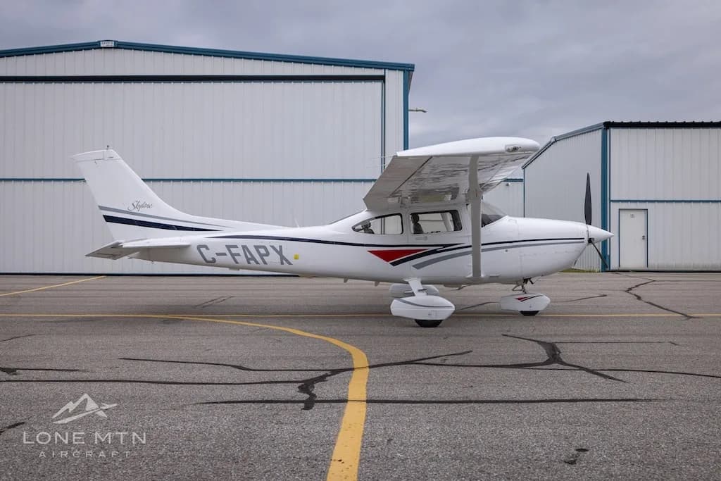 A white Cessna 182 Skylane with grey and blue stripes, registration C-FAPX, parked on an asphalt ramp in front of hangars.
