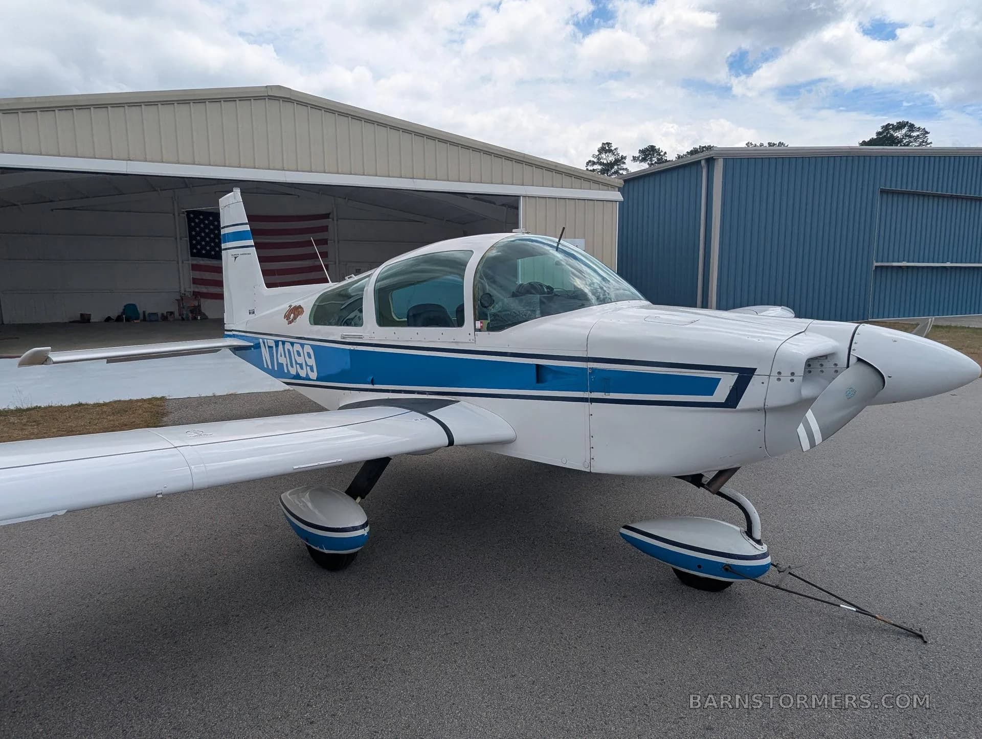 A white and blue Grumman AA-5 aircraft with registration N74099 parked in front of an open hangar with an American flag inside.