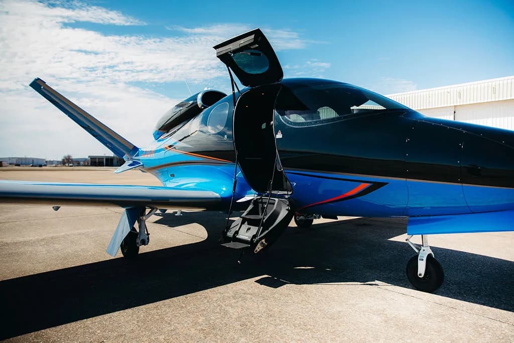 A close-up side view of a black and blue Cirrus Vision Jet (SF50) with its main entry door open on an airfield.