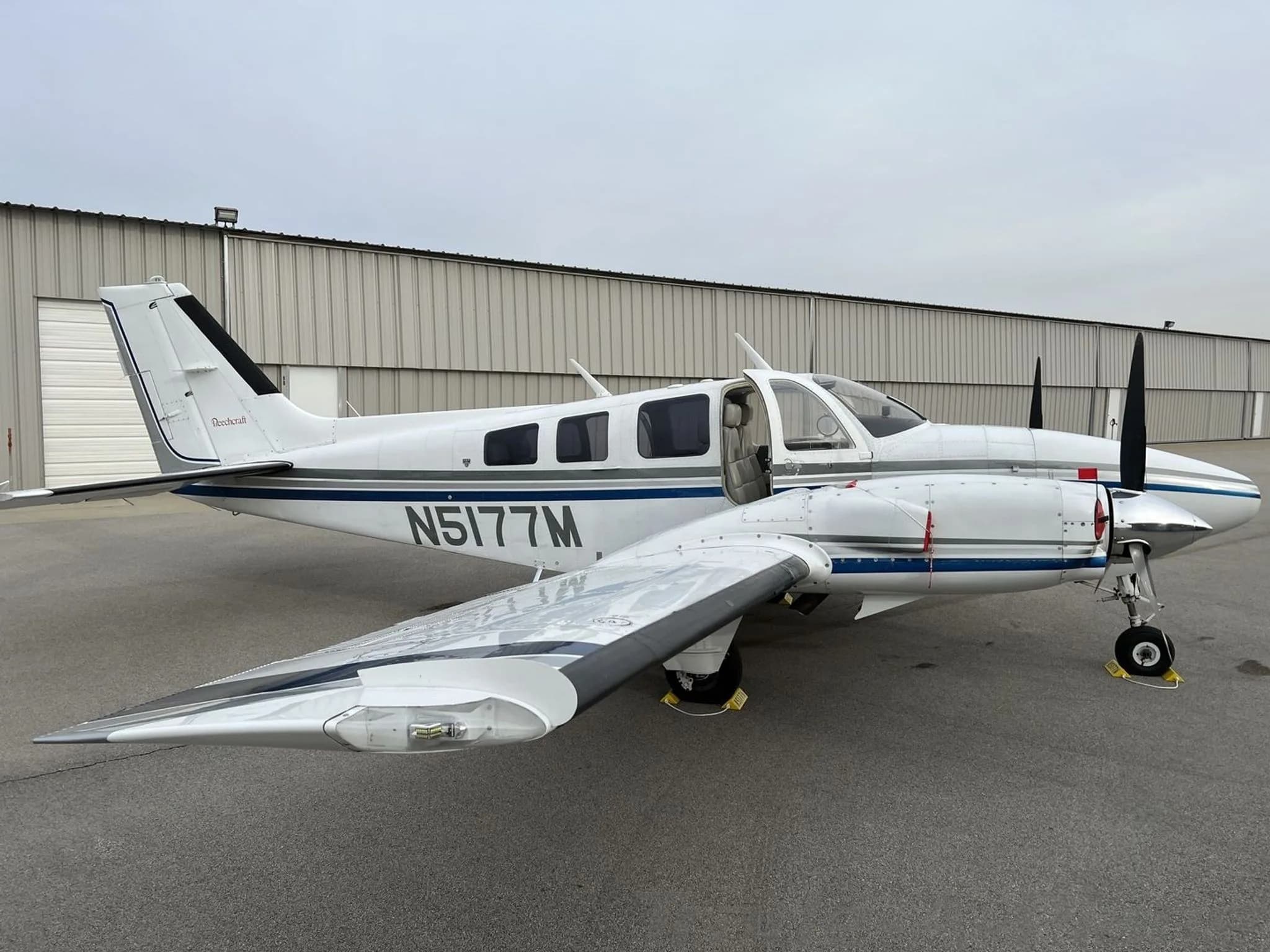 A three-quarter front view of the white and blue Beechcraft Baron N5177M with its main cabin door open, parked on an airfield.