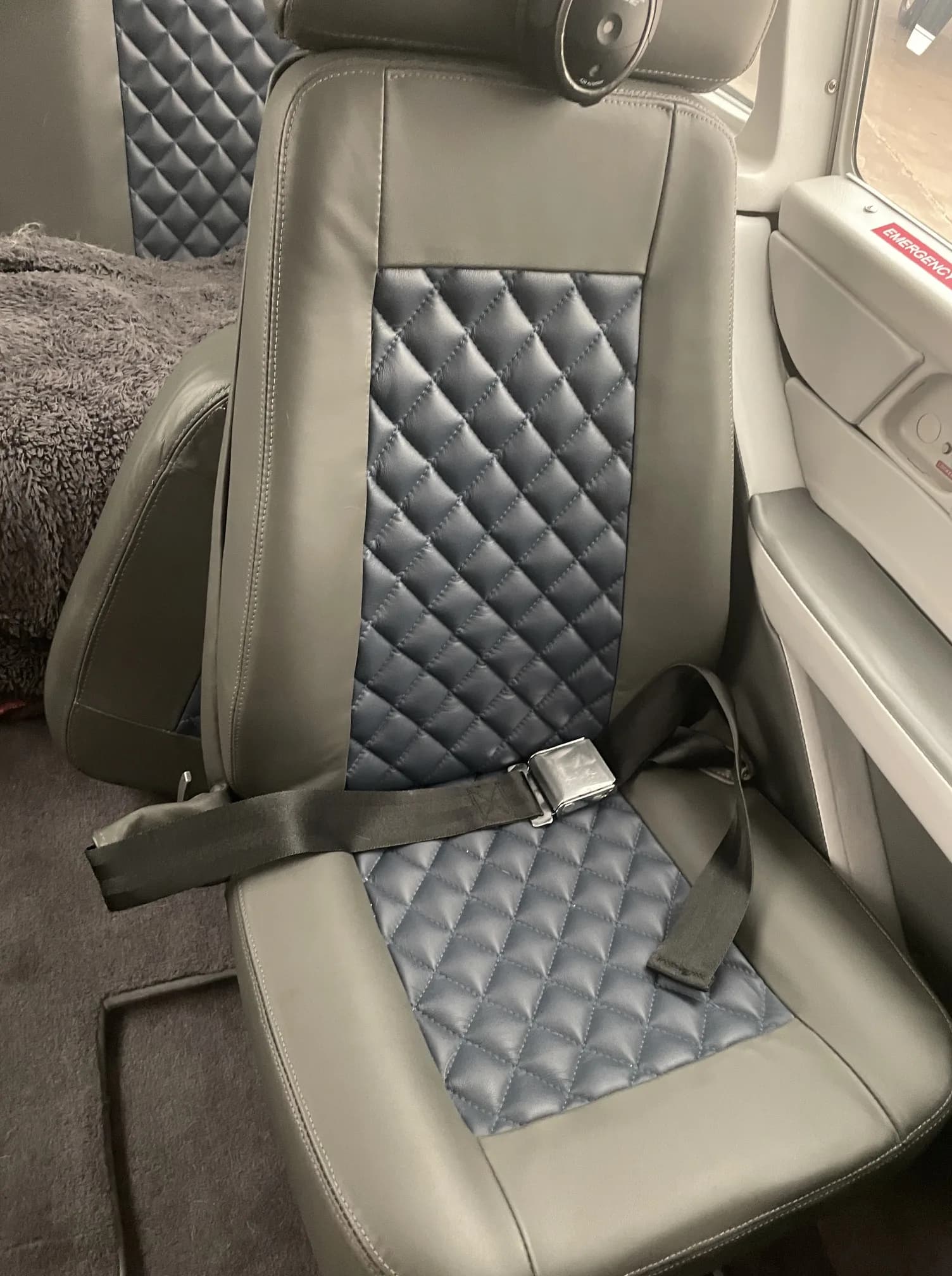 A close-up of a grey and blue quilted leather aircraft cabin seat with a seatbelt.