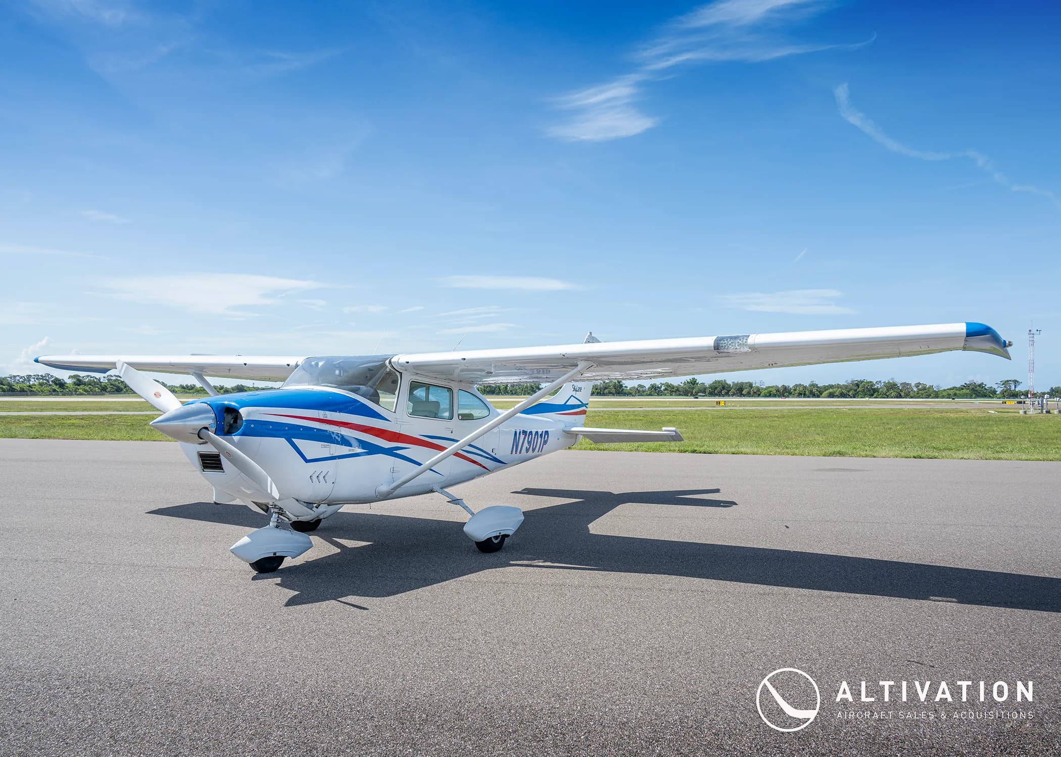 A front-quarter view of a white Cessna 182 Skylane with blue and red stripes parked on a tarmac.