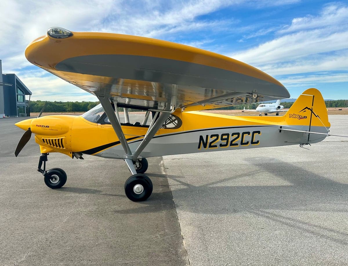 A yellow and gray NX Cub aircraft, N292CC, parked on an asphalt tarmac with a blue sky overhead.