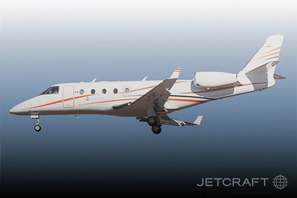 A white business jet with orange and black stripes in flight with landing gear extended.