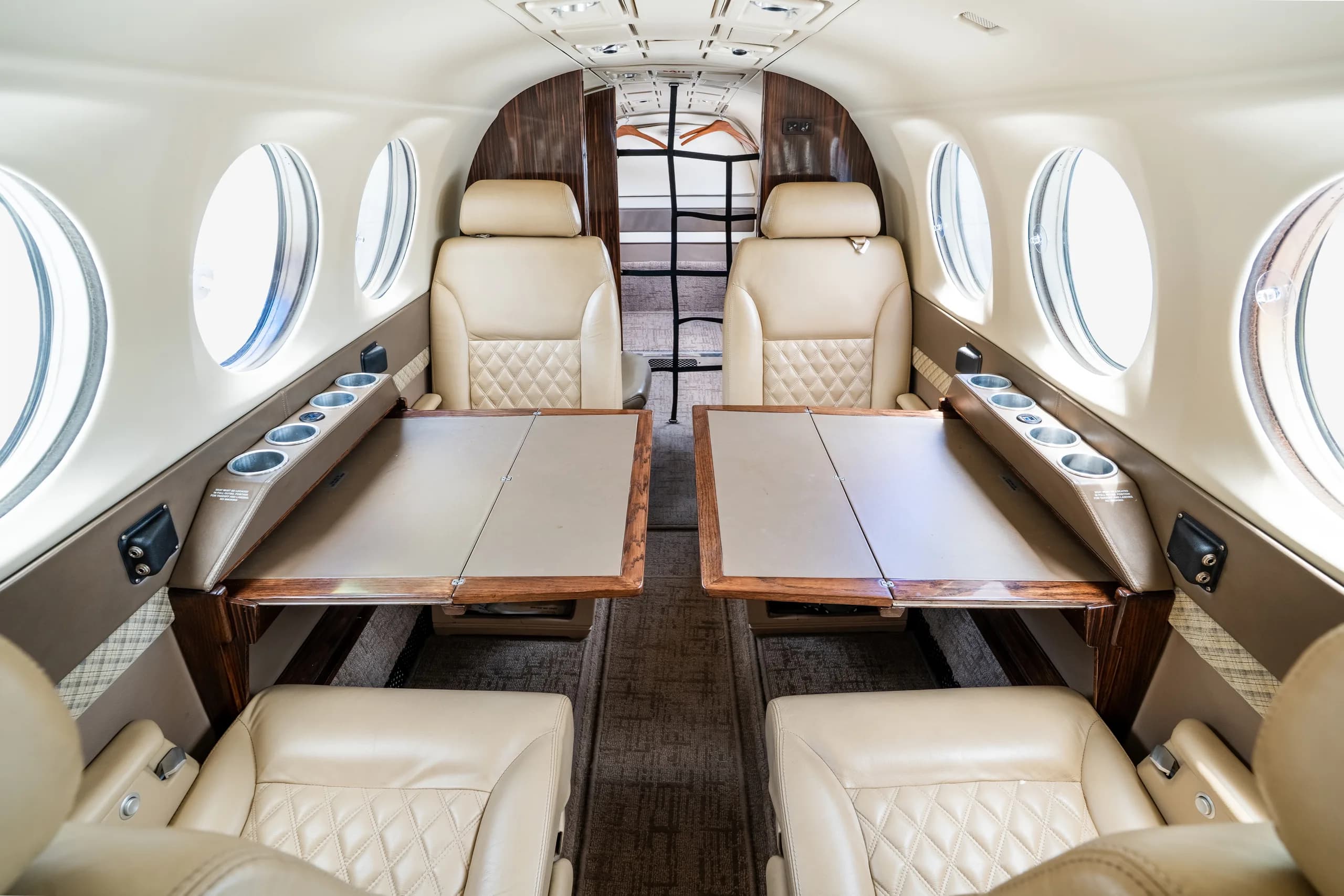The interior cabin of an aircraft featuring four beige leather seats with two fold-out tables.