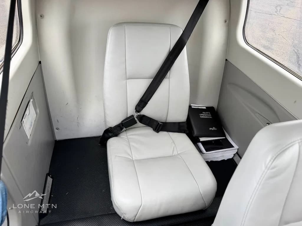 A view of the light gray rear cabin seat with a black seatbelt and a pilot's operating handbook on the floor.
