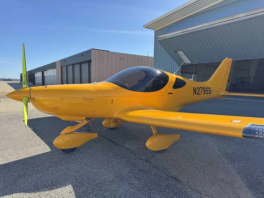 A bright yellow Bristell aircraft, registration N279SS, is parked on the tarmac with hangars in the background.
