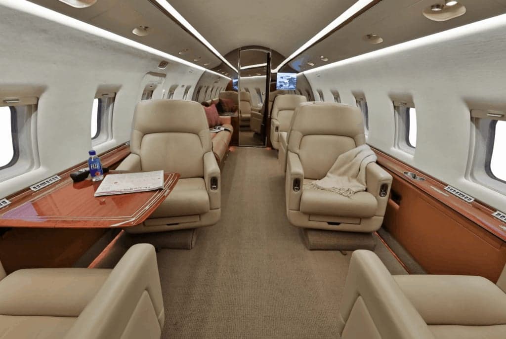 The elegant interior cabin of a business jet, with beige leather seating, a wooden table, and overhead lighting.