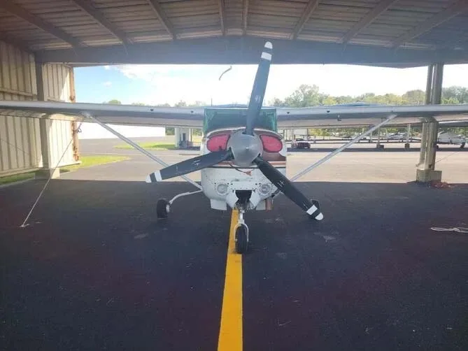 A front view of a white and brown Cessna Skylane RG II aircraft with a three-bladed propeller, parked under a hangar.