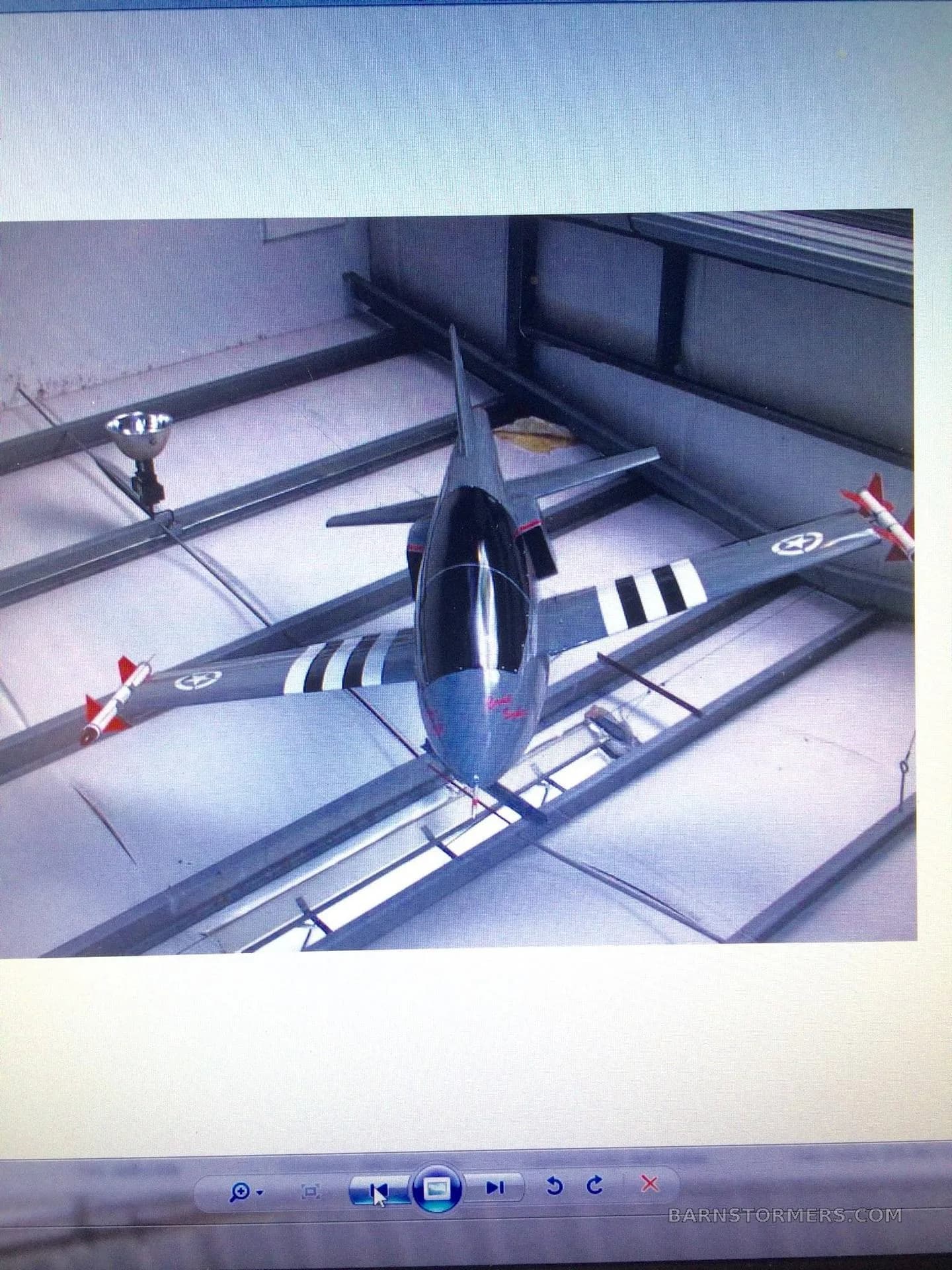 An overhead view of a grey model jet aircraft with black and white stripes and red missiles, displayed on a screen, hanging in a hangar.