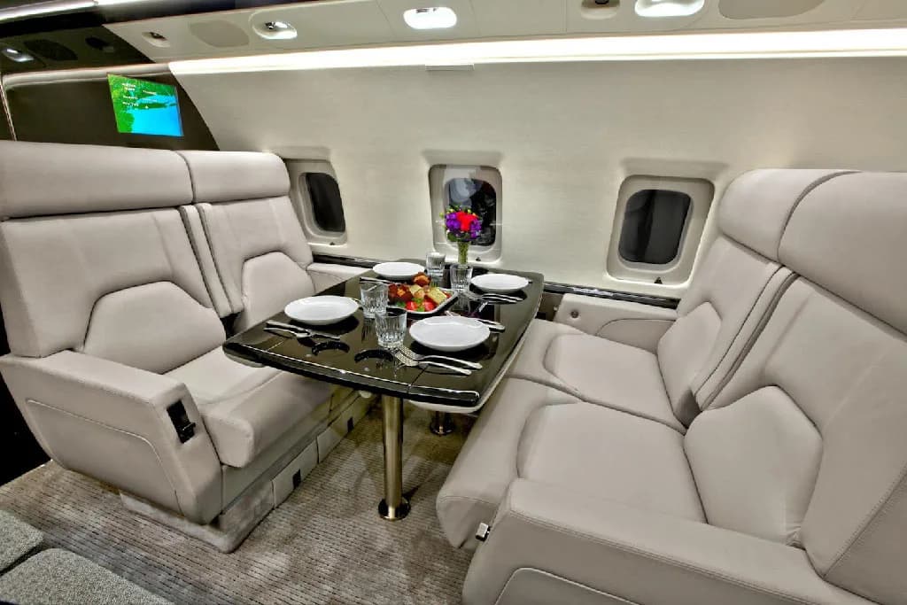 A section of the private jet cabin with two sets of facing light gray seats and a dark table set with plates and fruit.