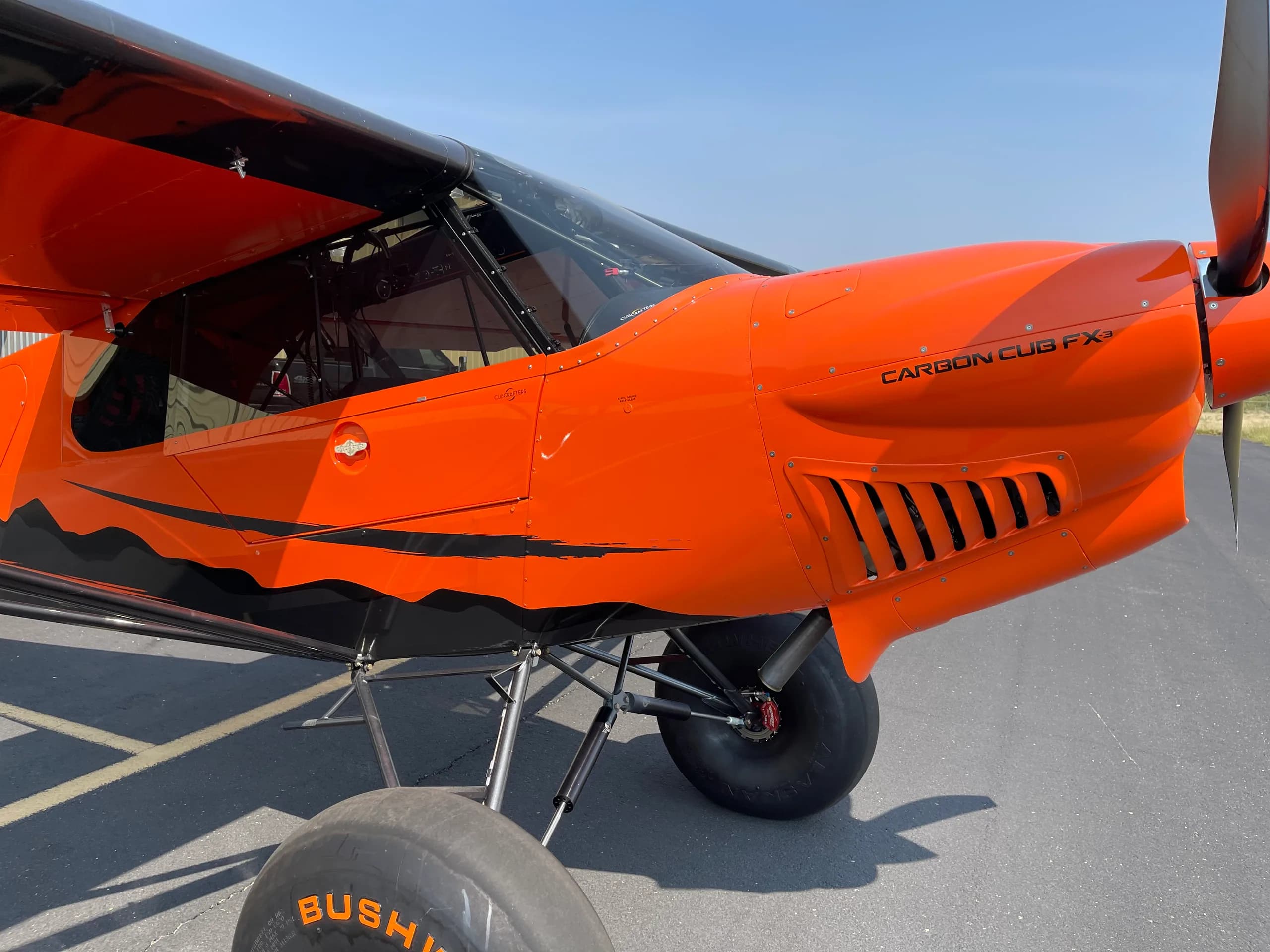 A close-up view of the front fuselage and engine cowling of the orange Carbon Cub FX-3 aircraft on an airport tarmac.
