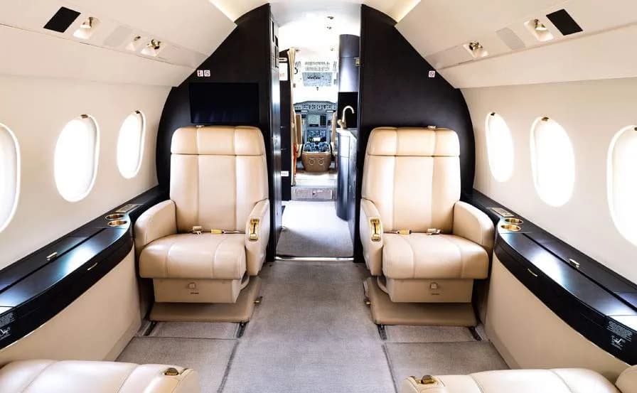 The interior cabin of a business jet, with beige leather seats and a view into the cockpit through an open door.