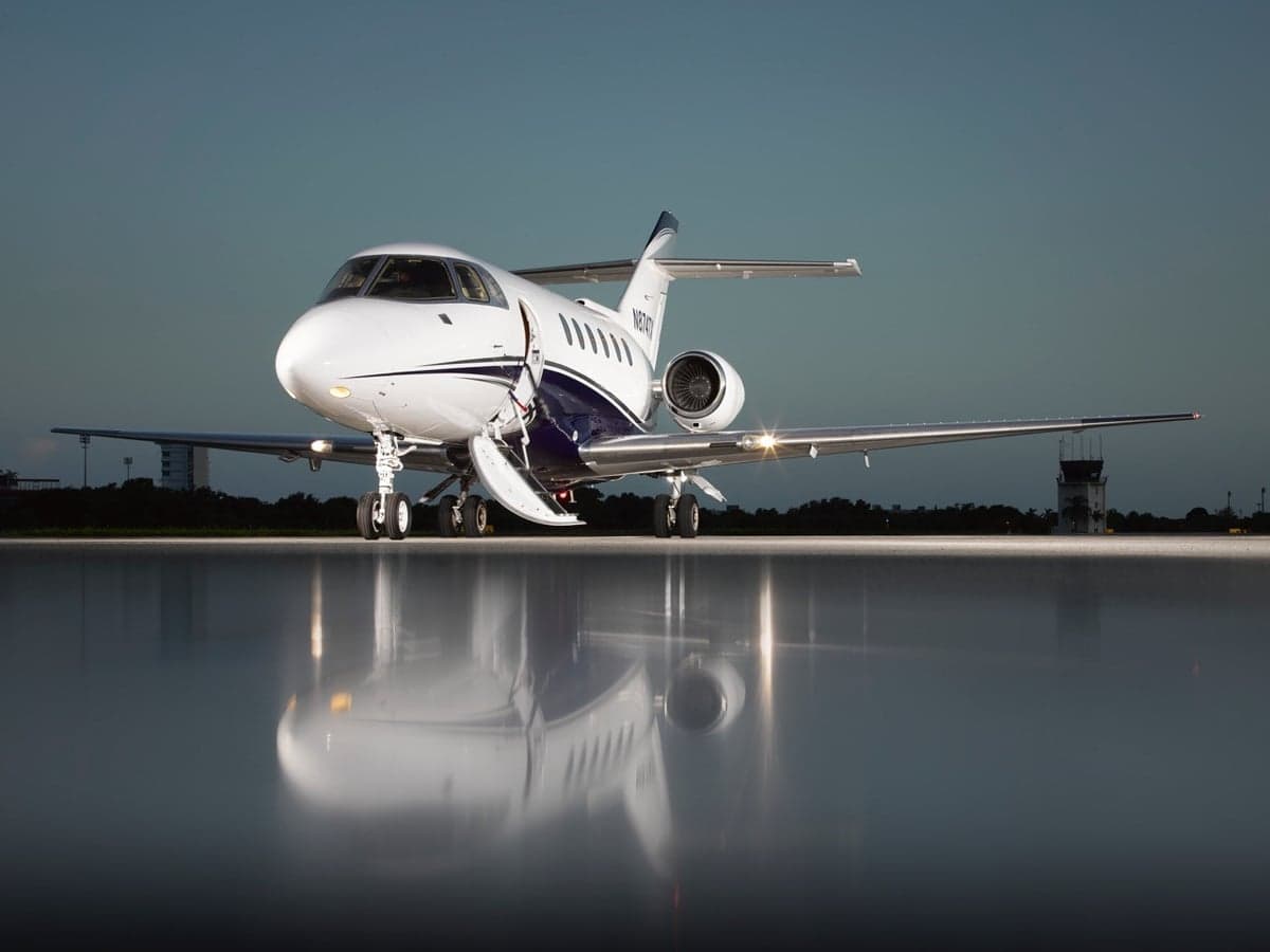 A white and blue business jet (N874TX) with its door open is parked on a wet tarmac, reflecting its image.