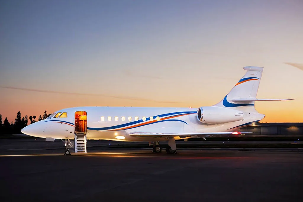 A white private jet with blue and orange stripes, parked on an airfield at dusk with its main door open and stairs extended.