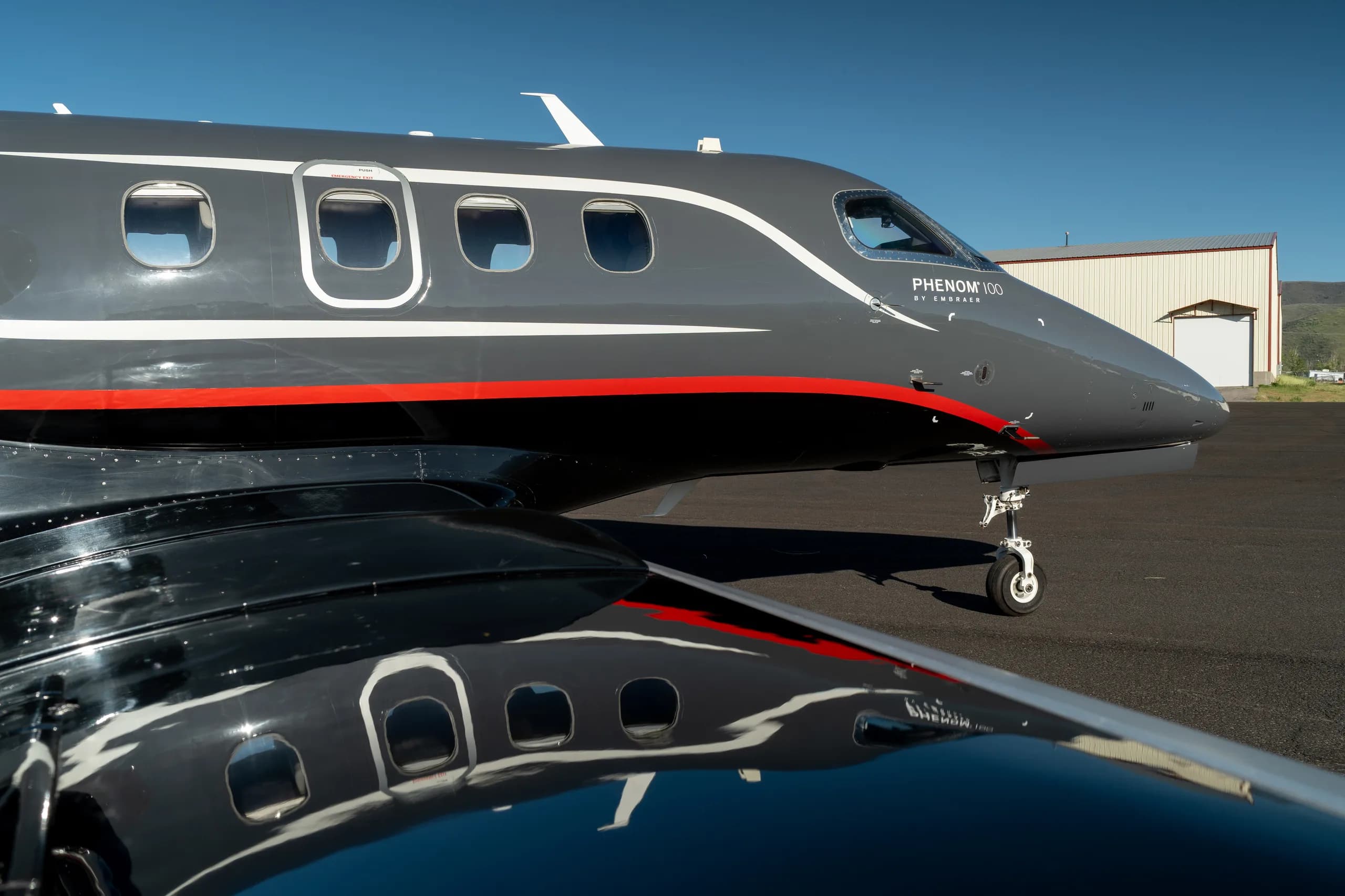 A close-up view of the front fuselage and wing of a dark gray Embraer Phenom 100 aircraft.