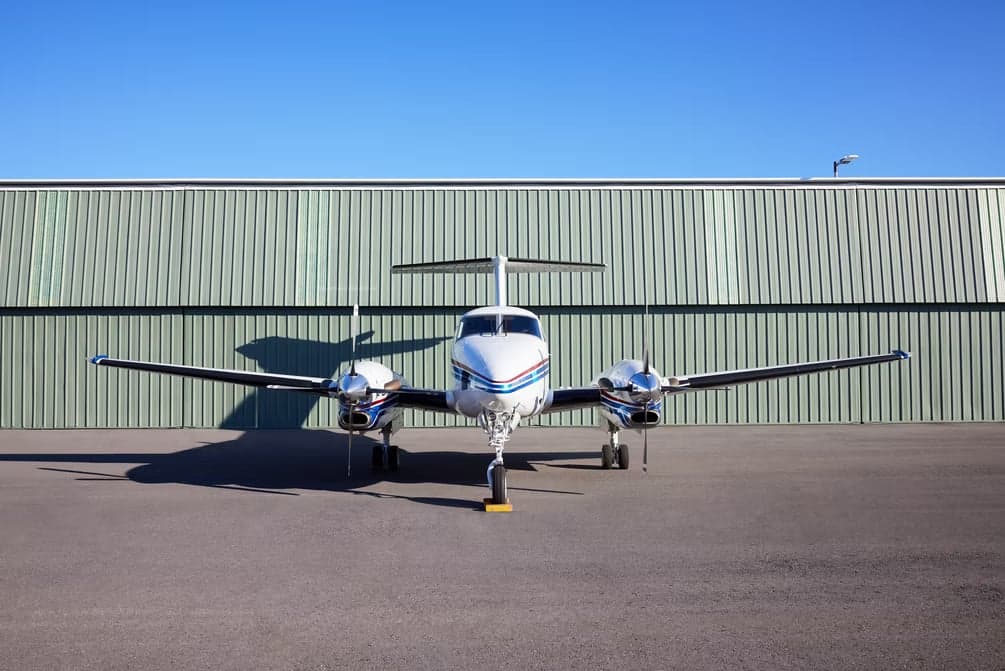 Front view of a Beechcraft King Air with a white and blue paint scheme parked in front of a hangar.