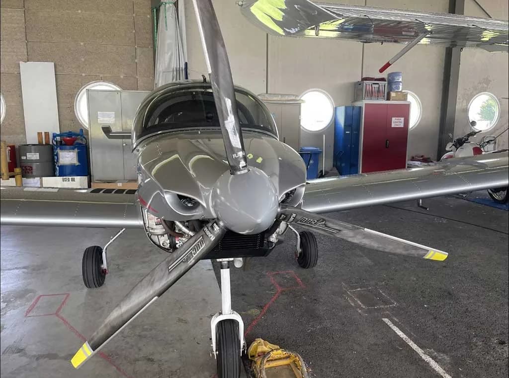 Head-on view of a silver Sonaca 200 aircraft with a three-bladed propeller, parked in a hangar.