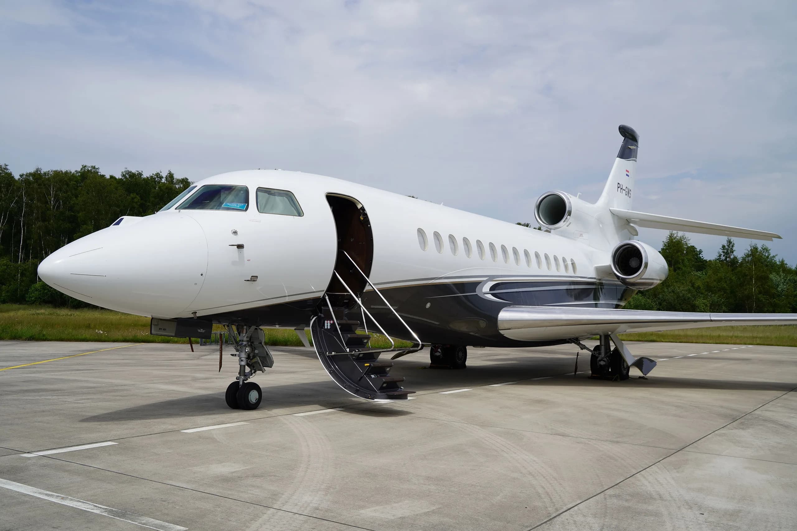 A white Dassault Falcon business jet with dark grey and silver accents is parked on an airfield with its main door open.