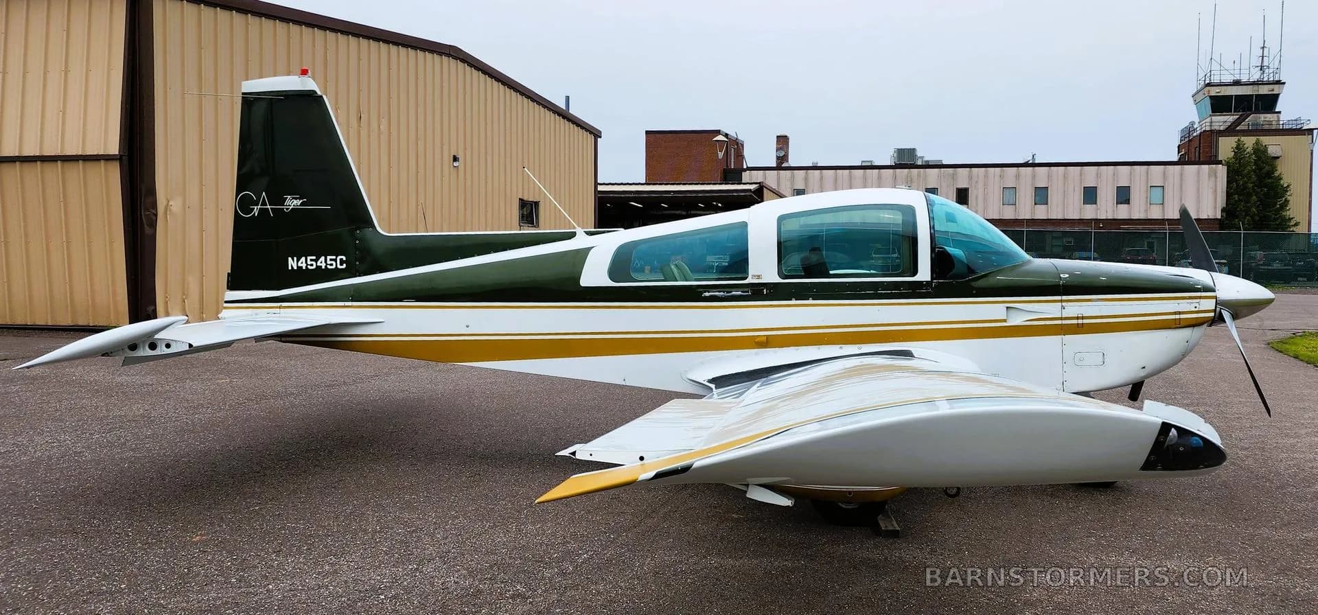 A side view of a white and dark green Grumman Tiger aircraft with gold stripes, parked on an asphalt ramp near a control tower.