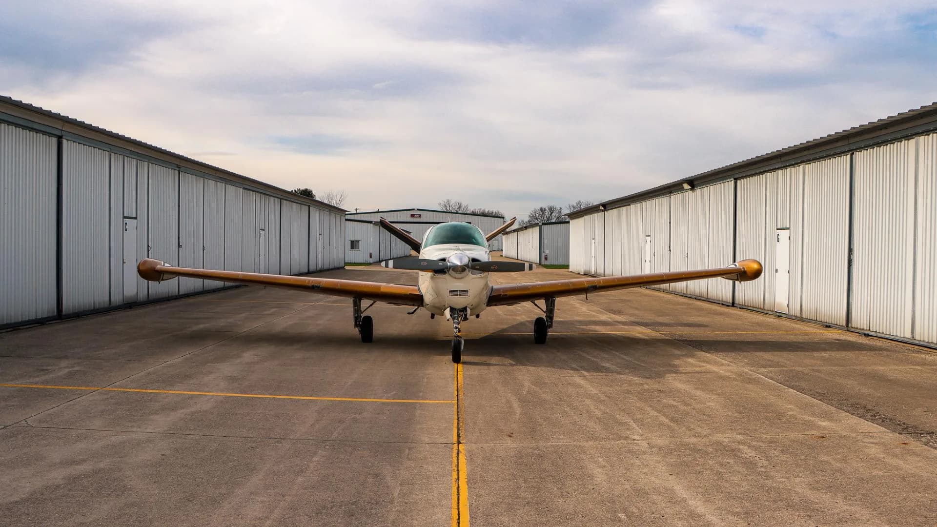 Front view of a white and gold Beechcraft Bonanza parked on an airfield between hangars.