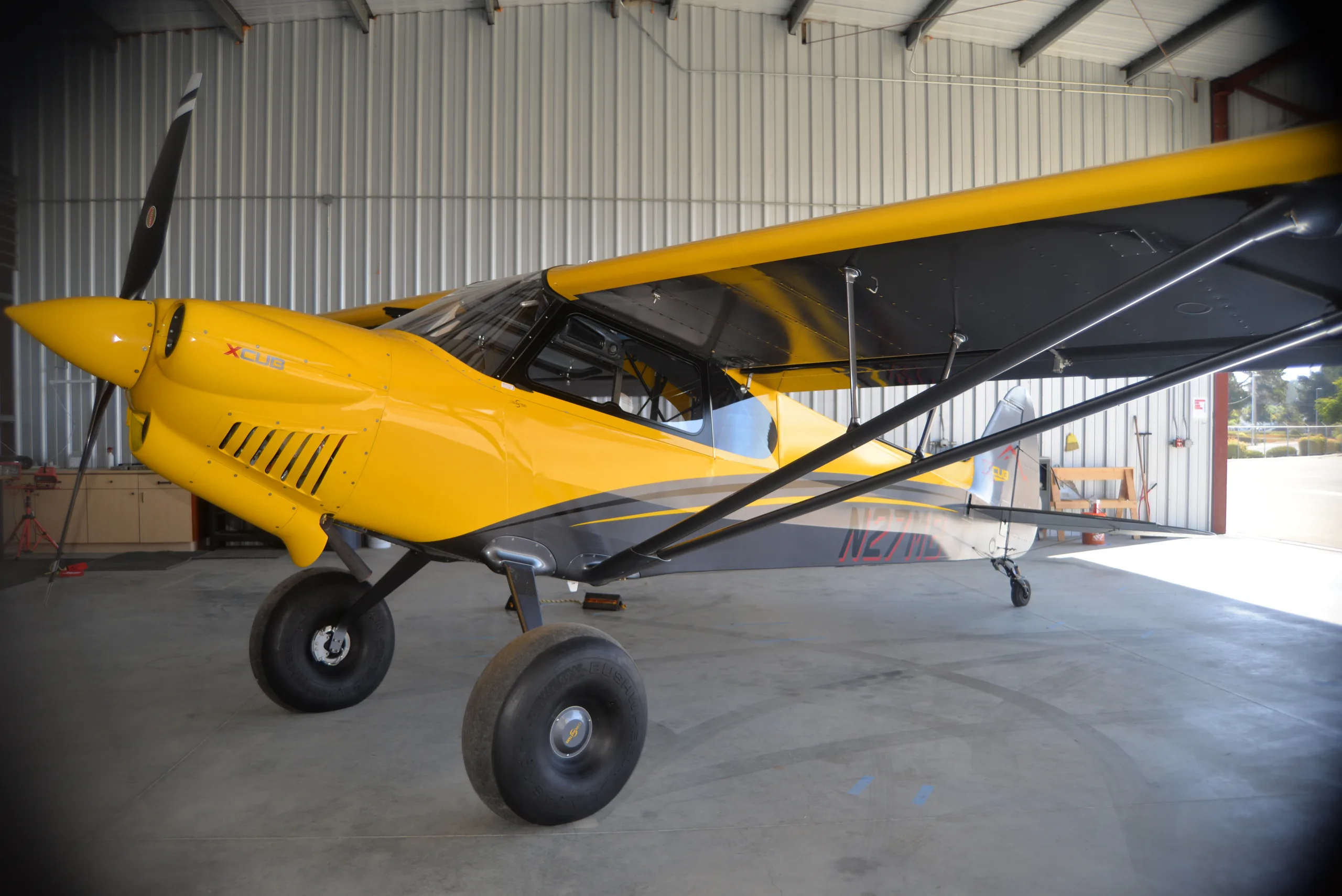 A yellow and gray CubCrafters XCub, registration N27MB, parked inside a hangar.