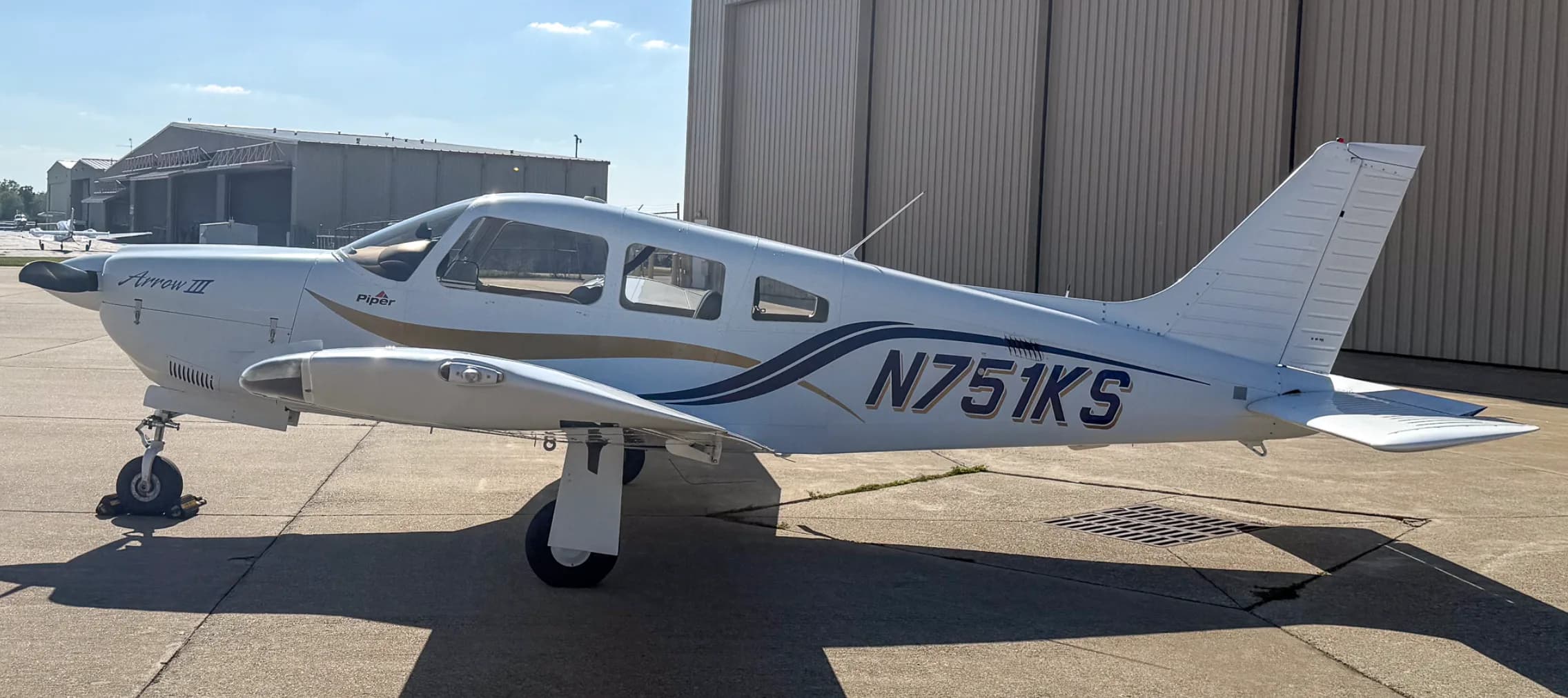 A left side view of the white Piper Arrow III aircraft, tail number N751KS, parked on an airport tarmac next to a hangar.